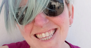 Headshot of C. Meaker, a queer white person with blue hair smiles at the camera. They wear a pink tank top on a sunny day with a white background.