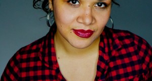 Headshot of Christin Eve Cato, an Afro-Latina woman smiling with red lips, a curly messy bun, and a red and black checkered shirt. 