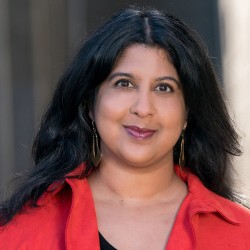 Headshot of Amrita Ramanan, wearing a red shirt with a black shirt underneath, smiling.
