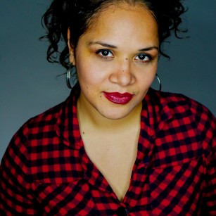 Headshot of Christin Eve Cato, an Afro-Latina woman smiling with red lips, a curly messy bun, and a red and black checkered shirt. 