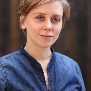 Headshot of Kate Tarker wearing a blue denim jumpsuit in front of a dark brown background